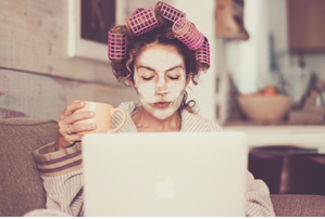 Woman in dressing gown sits at laptop with hair in rollers and a face pack on